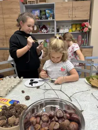 Miska pełna kasztanów i żołędzi stoi na stole, a w tle widać dzieci malujące farbami i ozdabiające orzeszki