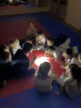 Dzieci leżą na materacach i słuchają z uwagą czytanej książki przy świetle latarki, w tajemniczej i magicznej atmosferze Nocy Bibliotek.