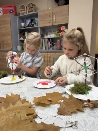 Dwie dziewczynki malują farbami, a na stole leżą liście i patyczki