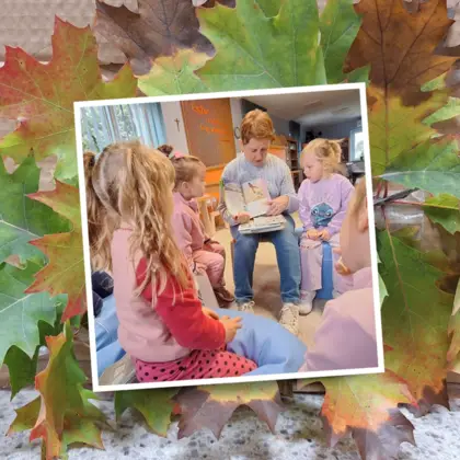 Zdjęcie przedstawiające dzieci stojące wokół kobiety siedzącej na krześle i pokazującej książki. Zdjęcie jest na zdjęciu z kolorowymi liśćmi.