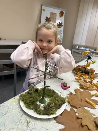 Uśmiechnięta dziewczynka trzyma rączki pod buzią i pokazuje swoje drzewo.