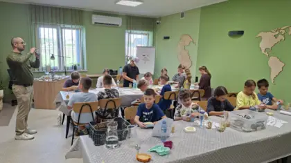 Dzieci przyglądają się skamieniałościom przy dwóch dużych stołach w sali, podczas gdy stojący po lewej stronie geolog wskazuje jedną z nich i o niej opowiada.