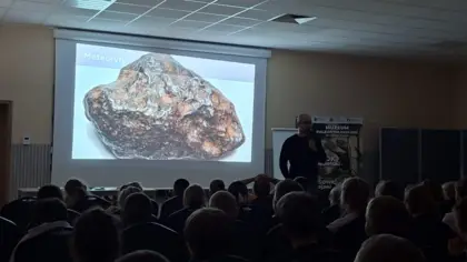 Mężczyzna opowiada o meteorycie widocznym w powiększeniu na ekranie, stojąc przed siedzącymi w zgaszonej sali dziećmi, ujęcie w mniejszym kadrze.