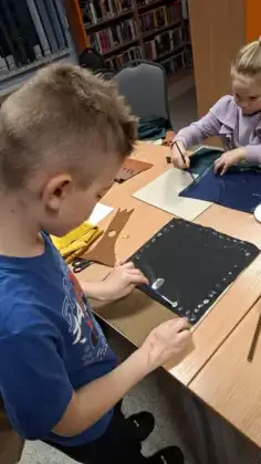 Dziewczynka i chłopiec siedzą przy stole i kleją materiał tapicerski.