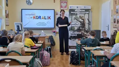 Zdjęcie wykonane od tyłu sali. Widzimy uczniów siedzących w ławkach tyłem do fotografa, a przed nimi prowadzącą warsztaty, która trzyma w dłoniach skrzynkę. W tle znajduje się baner Muzeum Paleontologicznego, a na tablicy wyświetlony jest temat zajęć „Zaczarowany koralik”.