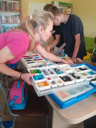 Pani instruktor układa na stoliku klocki lego w tle dwóch chłopaków składających konstrukcje.