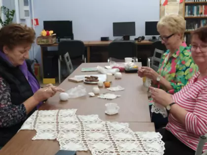 Panie siedzą przy stole w bibliotece i szydełkują.