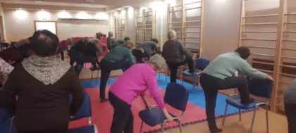 Seniorzy wykonują ćwiczenia relaksacyjne na sali ćwiczeń.