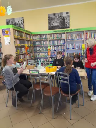 Uczestnicy zajęć siedzą przy stole w bibliotece i wykonują szopki bożonarodzeniowe.