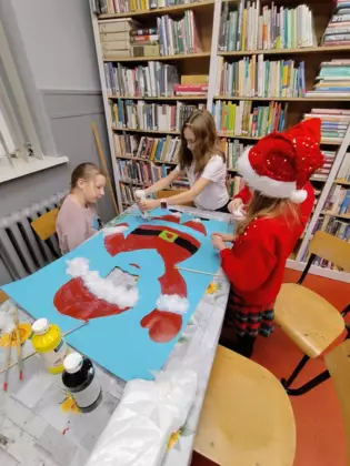 Dzieci we wnętrzu biblioteki wykańczają planszę na błękitnym brystolu z Mikołajkiem, którego twarz jest wycięta, aby dzieci mogły tam wystawić swoją twarz.