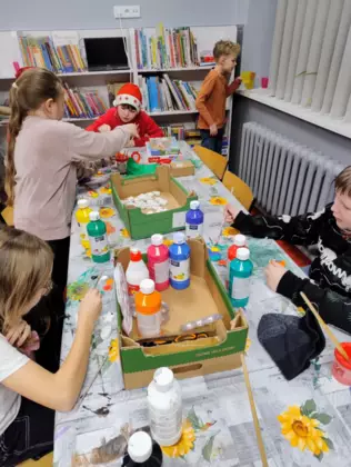 Dzieci we wnętrzu biblioteki pracujące przy długim stole, na którym stoją butelki z kolorowymi farbami.