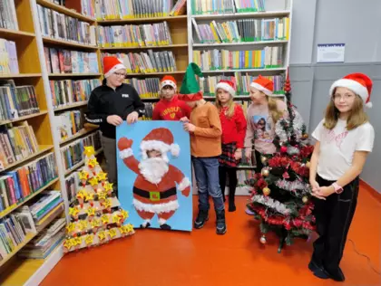 Zdjęcie z dziećmi pozującymi w grupie na tle regałów z książkami, w czerwonych czapkach krasnalowych. Przed dziećmi stoi plansza z Mikołajkiem i ubrana choinka.